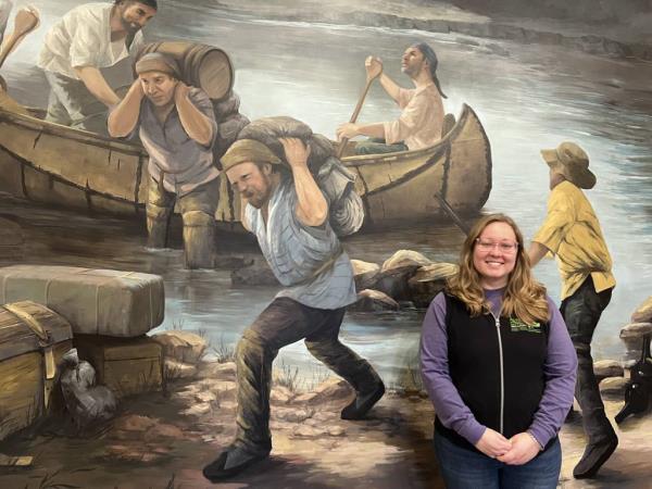 Jenna Krukowski, an interpretive naturalist with the Forest Preserve District of Will County, stands in front of a mural at the Isle a la Cache Museum in Romeoville that depicts French voyageurs and fur trappers in a scene harkening to 1750. 